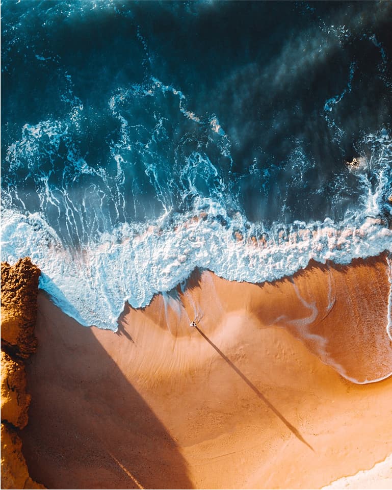 Aerial view of a beach with turquoise waves crashing onto golden sand, creating a stark contrast. A lone person walks along the shore.