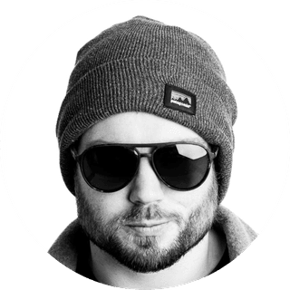 Man wearing sunglasses, a gray beanie, and a jacket, with a neutral expression, in a black and white portrait.