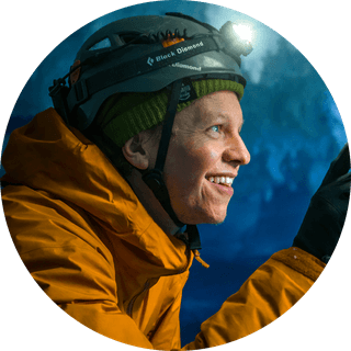 Person in an orange jacket and helmet with a headlamp, smiling in a dimly lit environment, possibly a cave or ice setting.