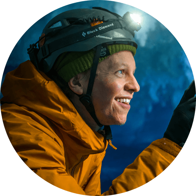 Person in an orange jacket and helmet with a headlamp, smiling in a dimly lit environment, possibly a cave or ice setting.